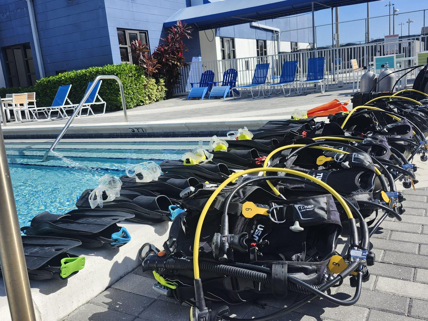 Scuba diving equipment lined up by a pool with blue building and chairs in the background