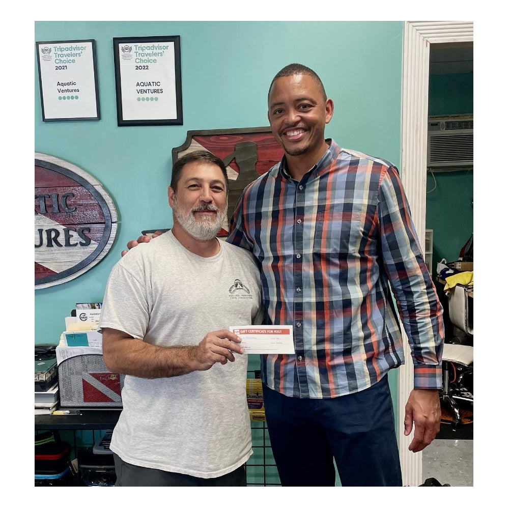 Two men standing together, one holding a check, in an office setting with framed documents on the wall.
