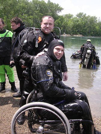 Person in a wheelchair with diving gear standing in water with other divers in the background.