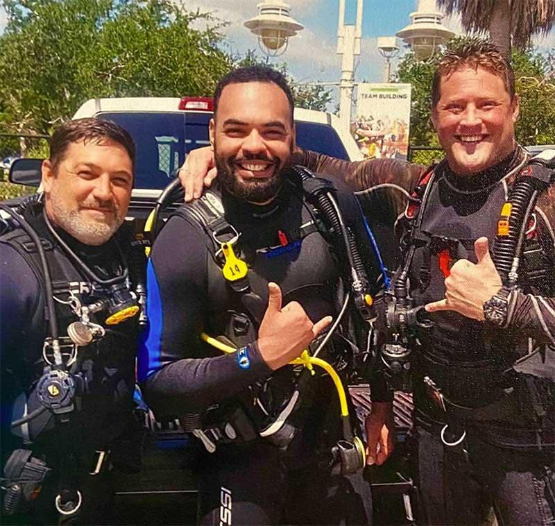 Three divers in scuba gear posing for a photo outdoors with a vehicle and trees in the background.